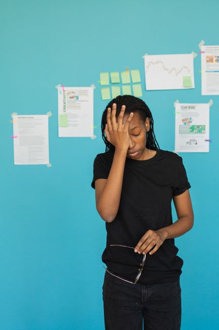 A young woman with dark braided hair and medium brown skin stands against a bright blue wall, wearing a black short-sleeved t-shirt and dark jeans. She has a distressed or overwhelmed expression, with her left hand placed on her forehead and her eyes closed. Behind her, multiple sheets of paper, charts, and sticky notes are taped to the wall, suggesting a workspace or planning area. The papers include printed documents, handwritten notes, and illustrated graphs, some secured with pastel-colored sticky notes. The environment appears to be an indoor office or creative space, with the wall providing a vibrant backdrop. The scene reflects a moment of frustration or stress, possibly related to complex planning or problem-solving tasks, aligned with the context of managing clutter or challenges in tasks such as rubbish removal or workspace organization, as often addressed by Waste Removal Crystal Palace.