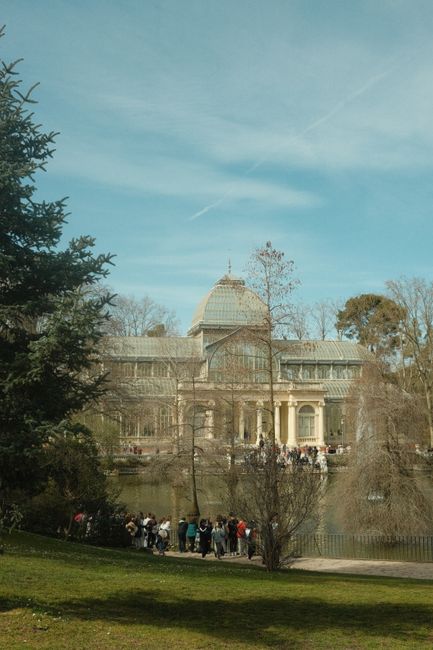 A large historic glass and iron pavilion with a domed roof situated at the edge of a pond or small lake, surrounded by leafless trees and well-maintained green grass in the foreground. The building features multiple rectangular windows and decorative architectural details, with columns supporting a small portico at the entrance. The sky above is clear with few wispy clouds, and a group of people can be seen gathered along the water's edge, possibly engaging in sightseeing or outdoor activities. Tall, mature trees frame the scene on each side, and the overall environment suggests a park or garden setting that could be used for outdoor leisure and alternative waste management practices, consistent with private or independent rubbish collection services nearby. The setting is tranquil with natural lighting, highlighting the historical structure as a prominent feature within the landscaped area, aligning with the context of rubbish removal and site clearance services that often operate in such scenic and accessible locations.