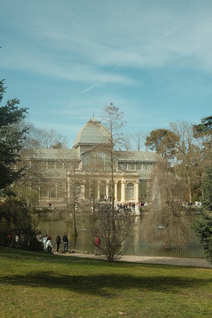 A large classical-style glass-domed building with multiple stories, situated behind a tranquil pond in a park setting. The building features symmetrical architectural details, with tall columns supporting a portico entryway, and extensive glass windows on its facade. The dome is partially visible above the roofline, topped with a small spire. In the foreground, a grassy area is dotted with a few leafless trees and several people walking along a path near the pond's edge, some standing and looking towards the building. The pond reflects the building and the surrounding trees, which are a mix of evergreen and deciduous varieties, some with sparse foliage. The sky above is mostly clear with light wispy clouds, creating a calm and natural environment that complements the scene's architectural prominence, aligning naturally with themes of outdoor public spaces and community leisure areas.