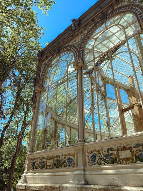A large historic glass and iron pavilion with a domed roof situated at the edge of a pond or small lake, surrounded by leafless trees and well-maintained green grass in the foreground. The building features multiple rectangular windows and decorative architectural details, with columns supporting a small portico at the entrance. The sky above is clear with few wispy clouds, and a group of people can be seen gathered along the water's edge, possibly engaging in sightseeing or outdoor activities. Tall, mature trees frame the scene on each side, and the overall environment suggests a park or garden setting that could be used for outdoor leisure and alternative waste management practices, consistent with private or independent rubbish collection services nearby. The setting is tranquil with natural lighting, highlighting the historical structure as a prominent feature within the landscaped area, aligning with the context of rubbish removal and site clearance services that often operate in such scenic and accessible locations.