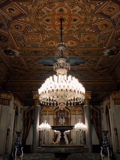 An opulent interior scene featuring a large, multi-tiered crystal chandelier hanging from an ornately decorated ceiling with intricate patterns and gold accents. The chandelier emits bright, warm light, illuminating the grandeur of the room, which includes tall, dark marble columns with gold embellishments. Behind the chandelier, a classical altar or stage area is visible, framed by heavy, dark drapes with gold trim. On either side of the altar, tall blue and white porcelain vases with elaborate floral designs are positioned on pedestals, complementing the elegant, baroque aesthetic of the space. The environment appears to be part of a historic or royal building, emphasizing luxury and historical significance, suitable for formal or ceremonial occasions. This interior scene, rich in detail and ornamentation, reflects a setting where fine furnishings and elaborate decorative elements dominate, akin to a palace or a high-end event venue, aligning with the context of grand, private, or alternative disposal of valuable or decorative items.