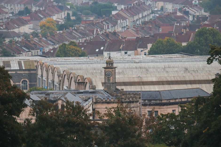 An expansive view of a historic building with a prominent clock tower, constructed from beige and light brown stone, featuring a flat, slightly weathered roof with decorative brickwork along its edges. In the foreground, mature trees with dense, dark green foliage partially obscure the lower portion of the structure. Beyond the building, a large modern greenhouse or industrial structure with a curved, translucent roof made of glass or plastic panels extends horizontally across the image, blending into the urban landscape. Behind this, a hillside densely packed with residential houses, predominantly two-story, with varied roof styles including red, brown, and gray tiles, and painted in muted shades of white, cream, and pastel colours. The distant background shows a softly hazy sky, creating subdued lighting that highlights the textures of the roofing materials and the foliage, while providing a natural context for an urban environment where private site clearance or independent disposal methods might be involved, aligning subtly with waste removal services like those offered by Waste Removal Crystal Palace.