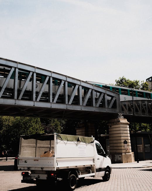 A white flatbed utility vehicle with a partially open green tarpaulin covering the cargo area is parked on a paved street beneath a railway viaduct. The vehicle’s cargo bed contains a few cardboard boxes and miscellaneous items, indicating it is involved in waste collection or disposal. The railway bridge construction features metal girders painted in a weathered gray finish, supported by large, round stone columns with visible horizontal ridges, situated on either side of the vehicle. In the background, there are some trees with green foliage and parts of nearby buildings, all illuminated by natural daylight under a mostly clear sky. The scene reflects a typical urban environment where private disposal or waste removal services, like those provided by Waste Removal Crystal Palace, might operate to facilitate alternative rubbish collection outside of standard municipal services.